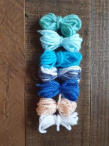 A photo of seven small yarn bundles to show the colours that will be used in a future version of the wreath, representing frost and winter, which are white, peach, royal blue, variegated blues & whites. cyan, baby blue and teal