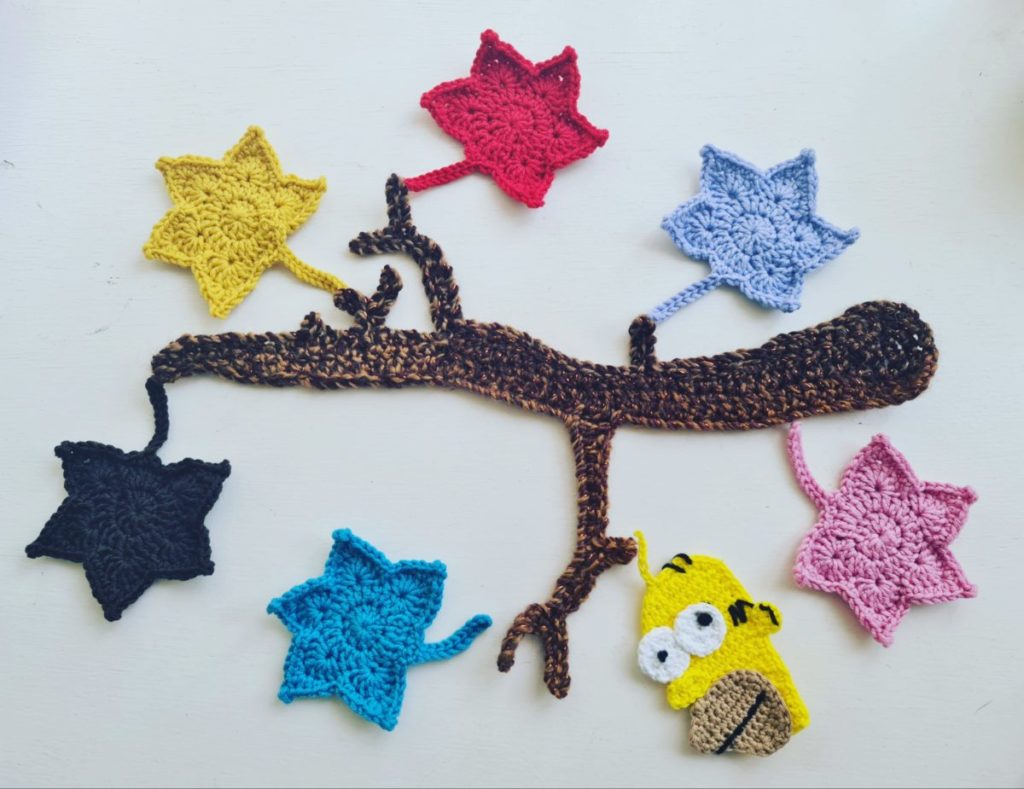 A flat piece of crochet that depicts a brown tree branch, with six different coloured leaves hanging from the branch. On one of the branches a crochet emblem of the cartoon character Homer Simpson is hanging in place of a leaf. The image is called 'the wrong kind of leaf'