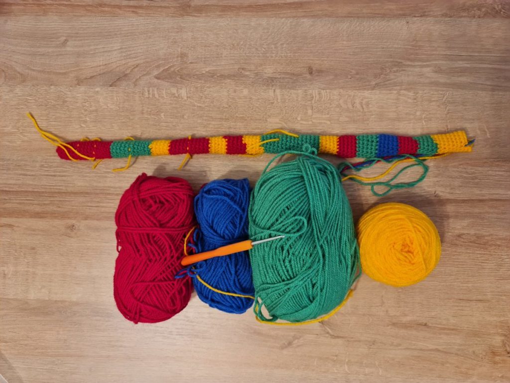 A long tube of crochet made up of different sized stripes of wool coloured red, blue, green and yellow. The picture shows the tube alongside four balls of wool and a crochet hook.
