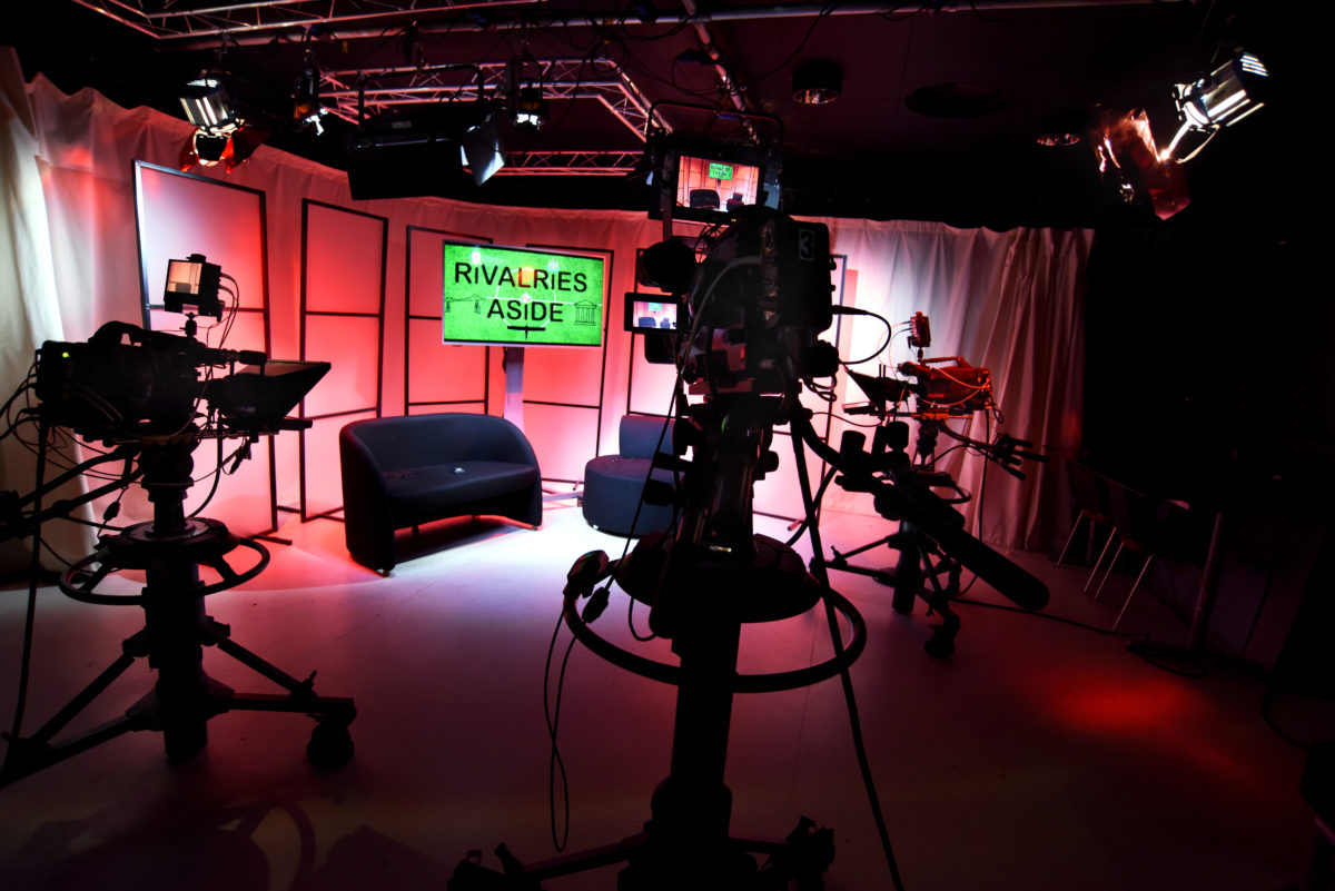 A professional newsroom and studio with recording equipment visible behind the scenes.