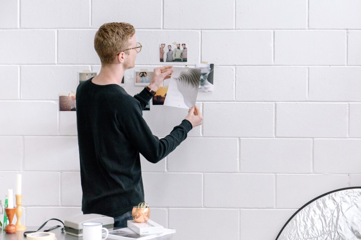 Man decorating his room by hanging pictures up on the white, brick wall