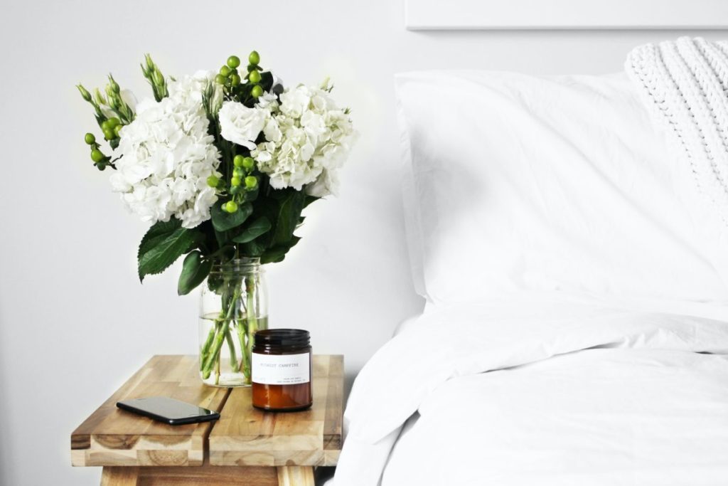 Image of a soothing bedroom, with white bedsheets and a vase of white flowers.