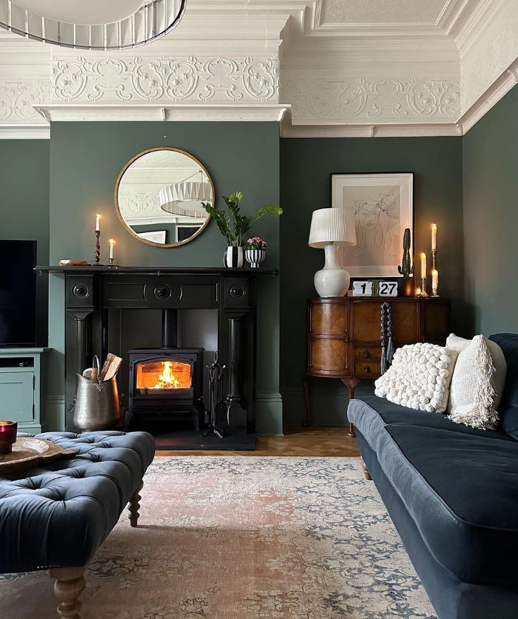 Image shows lounge painted in Farrow and Ball's Green Smoke paint, with a navy blue sofa and log burning stove.