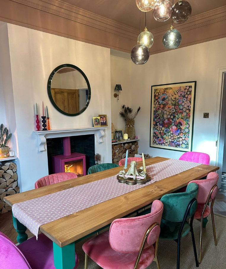 Casa Oldcorn dining room features a bright pink log burner, rustic wood dining table and pink and green velvet dining chairs.