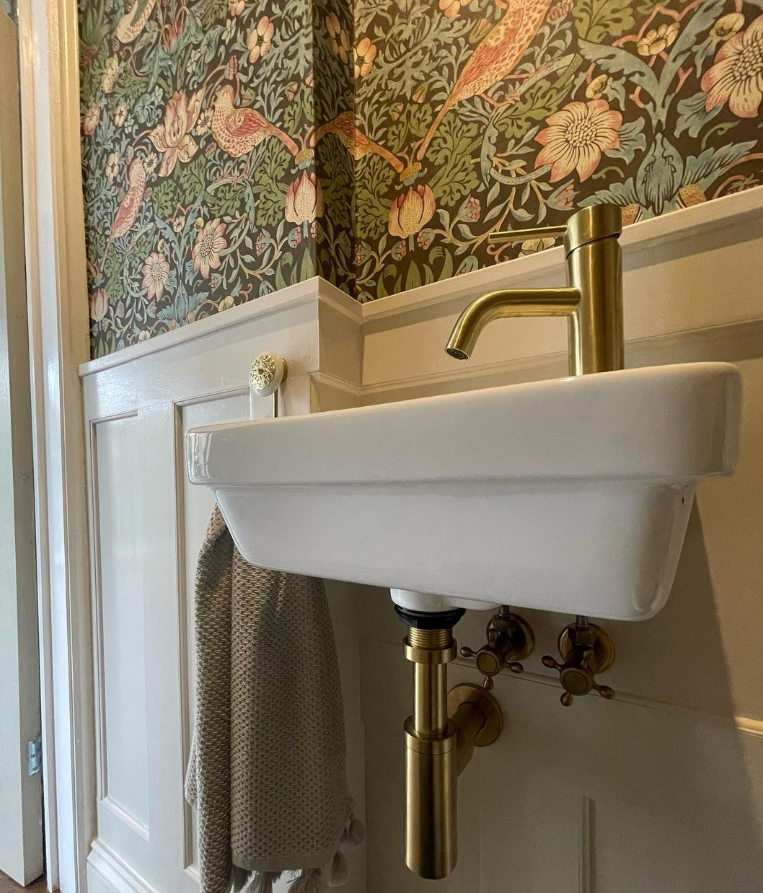 Image shows a white basin, gold tap and William Morris Strawberry Thief wallpaper.