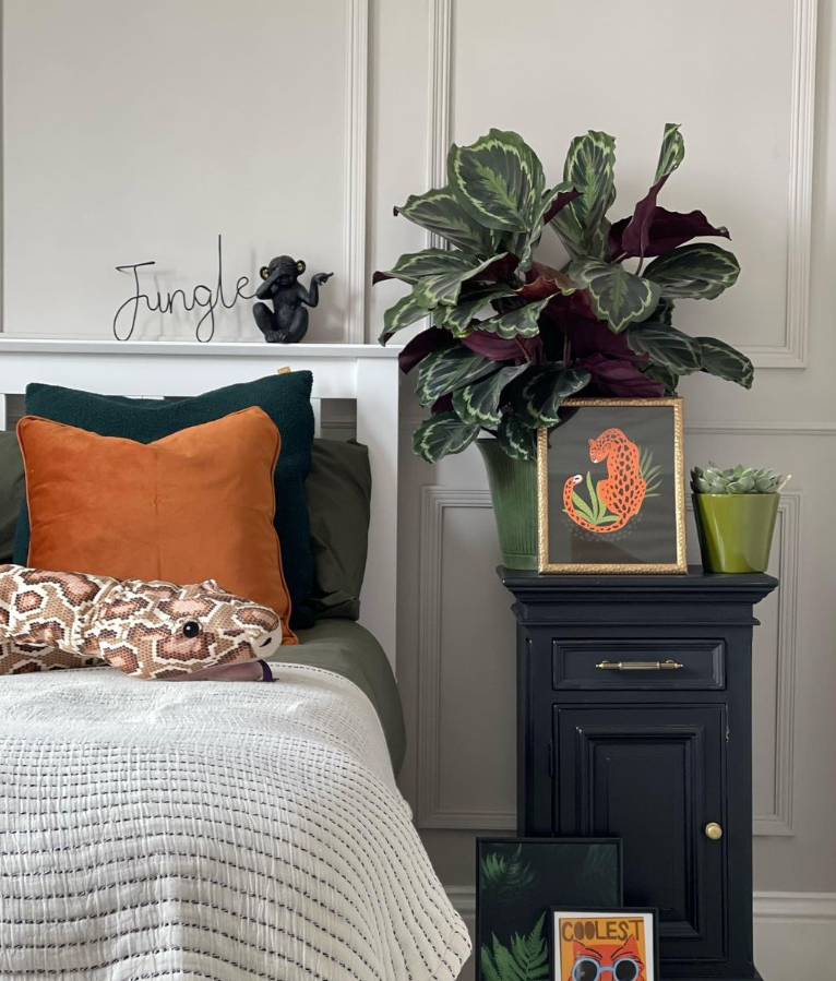 Image shows part of a bed with white bedding, black and orange cushions, panelled walls, houseplants and prints.