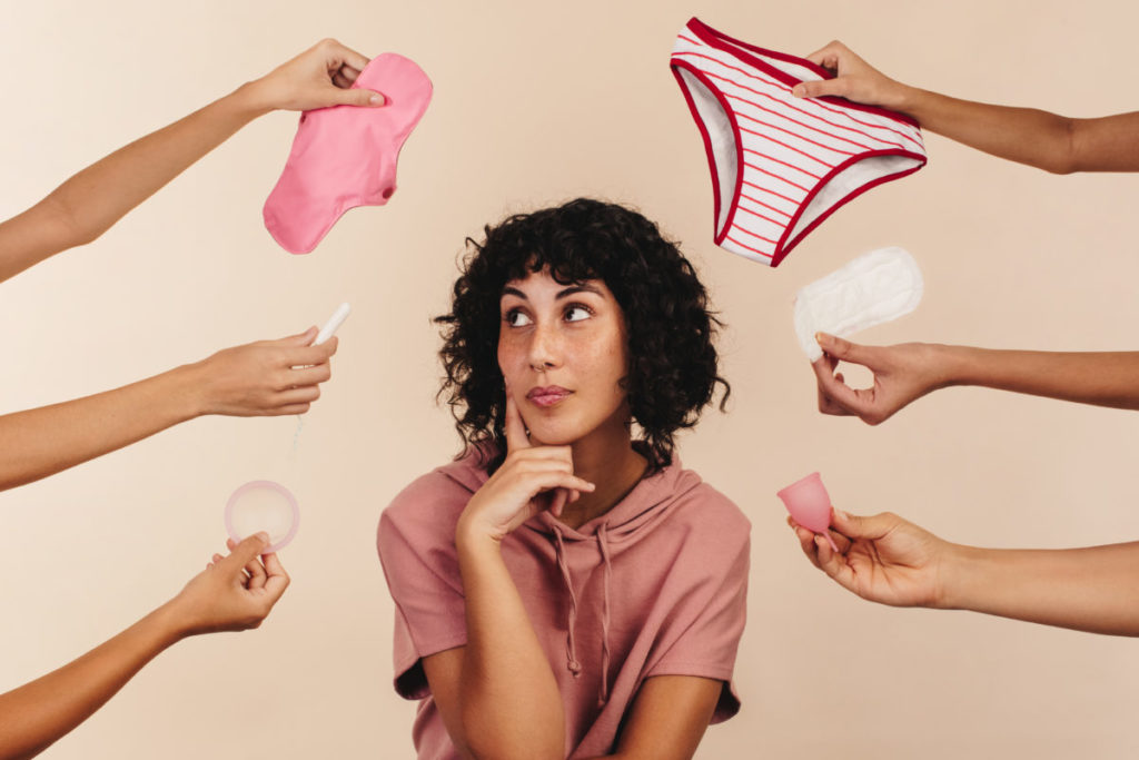 Thoughtful young woman looking away while surrounded by hands holding different reusable and non-reusable sanitary products.