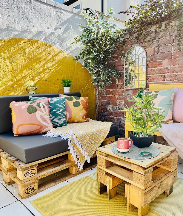 Image shows a yarden with a yellow and white painted brick wall, pallet seating and soft furnishings.