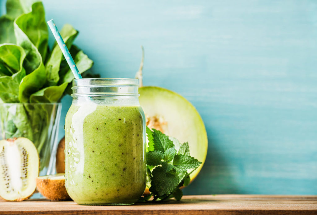 Freshly blended green fruit smoothie in glass jar with straw. Turquoise blue background