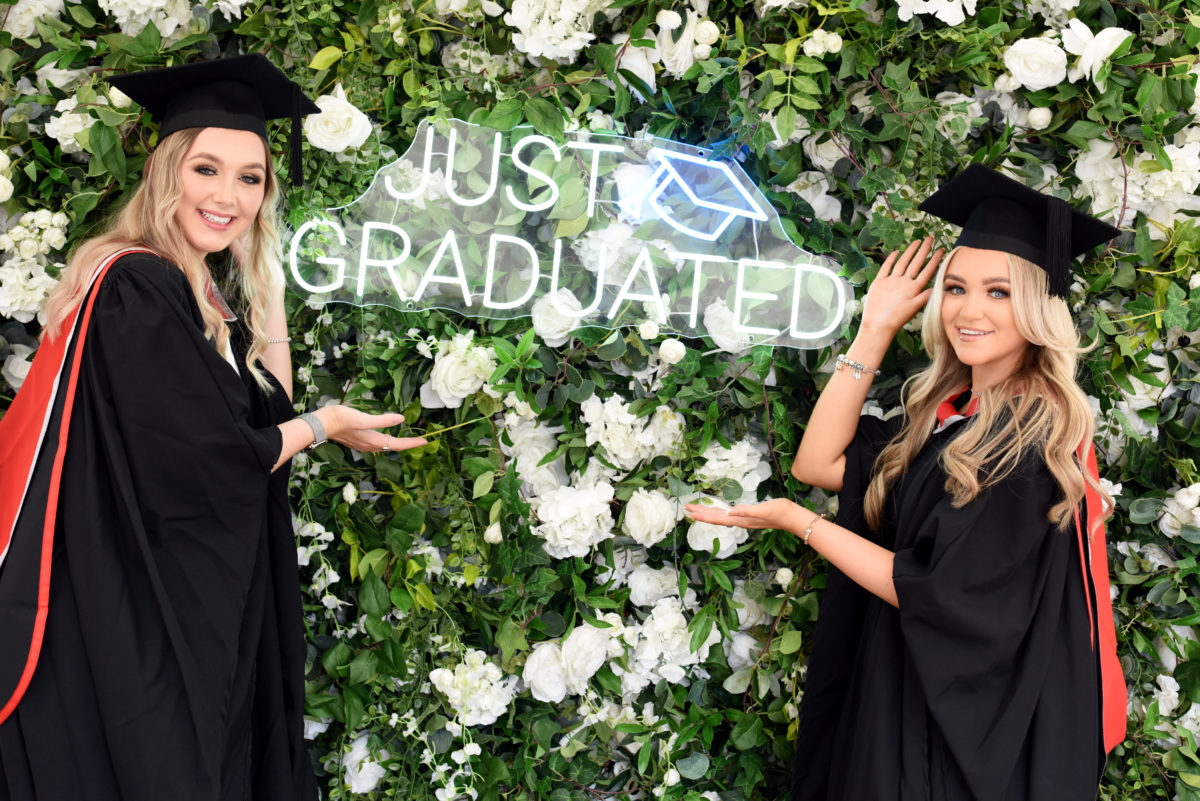 Two graduates posing with the flower wall and a neon sign saying 'just graduated' 
