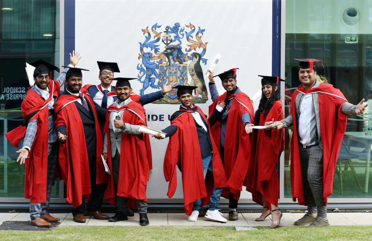 A large group of master's graduates standing in front of the Teesside University crest