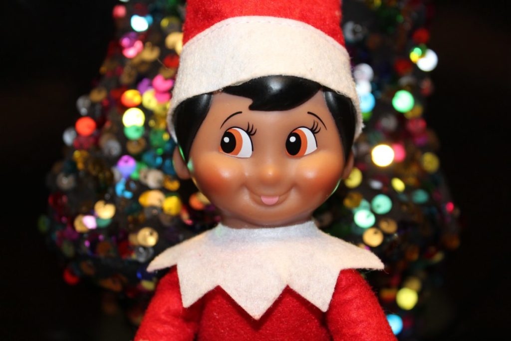 Naughty elf doll pictured smiling in front of a Christmas tree