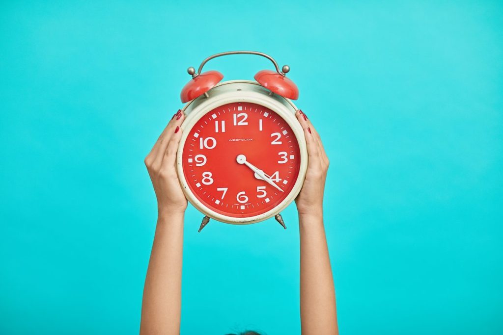 A woman holding up a big red alarm clock against a turquoise background.
