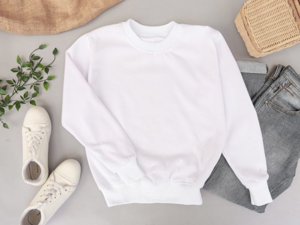 Flat lay image of an outfit including white plimsolls, white sweatshirt, blue jeans and foliage. 