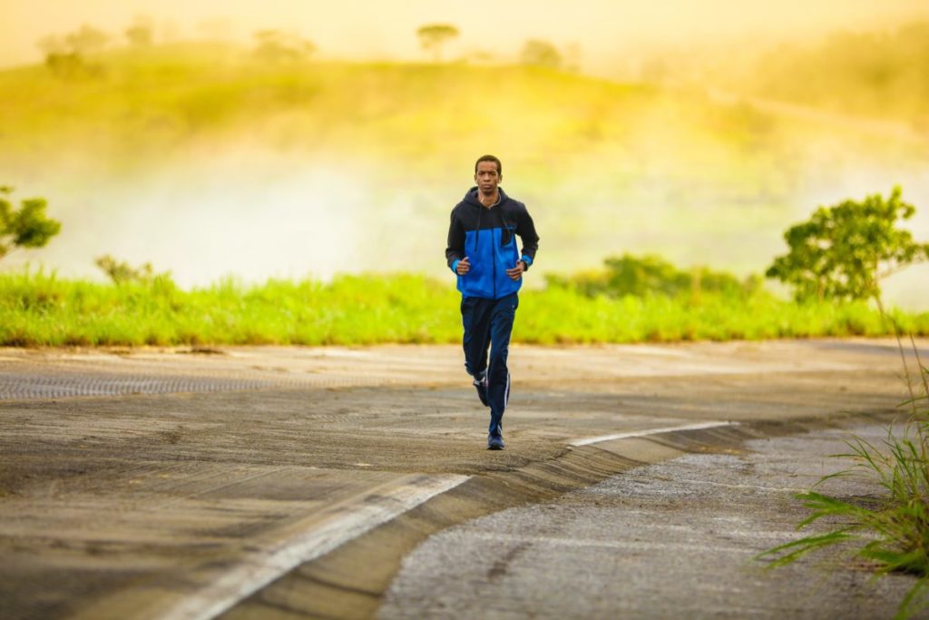 A man running down a road during the day, wearing a blue hoodie and training bottoms.