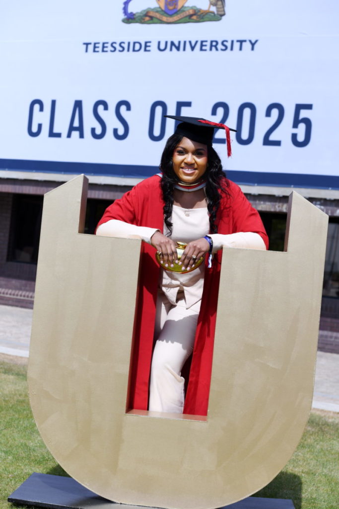 Graduate leaning on a giant, gold Teesside University logo
